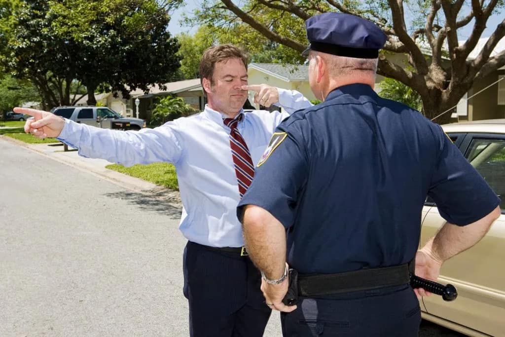 A man in a shirt and tie stands on a residential street with his arms outstretched, touching his nose, while a police officer observes—sobriety tests like this can help prevent car accidents and may require the guidance of lawyers.