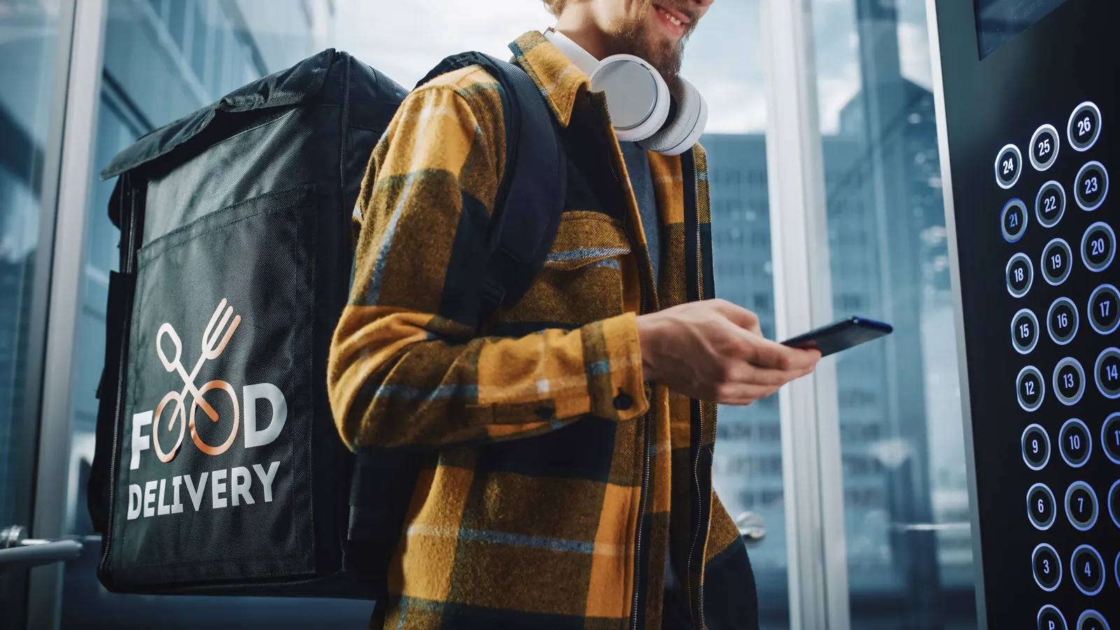 A smiling food delivery worker wearing a yellow plaid shirt, headphones, and a large backpack enters an elevator while checking a smartphone. The backpack has a "Food Delivery" logo on it. Elevator buttons are visible on the right.