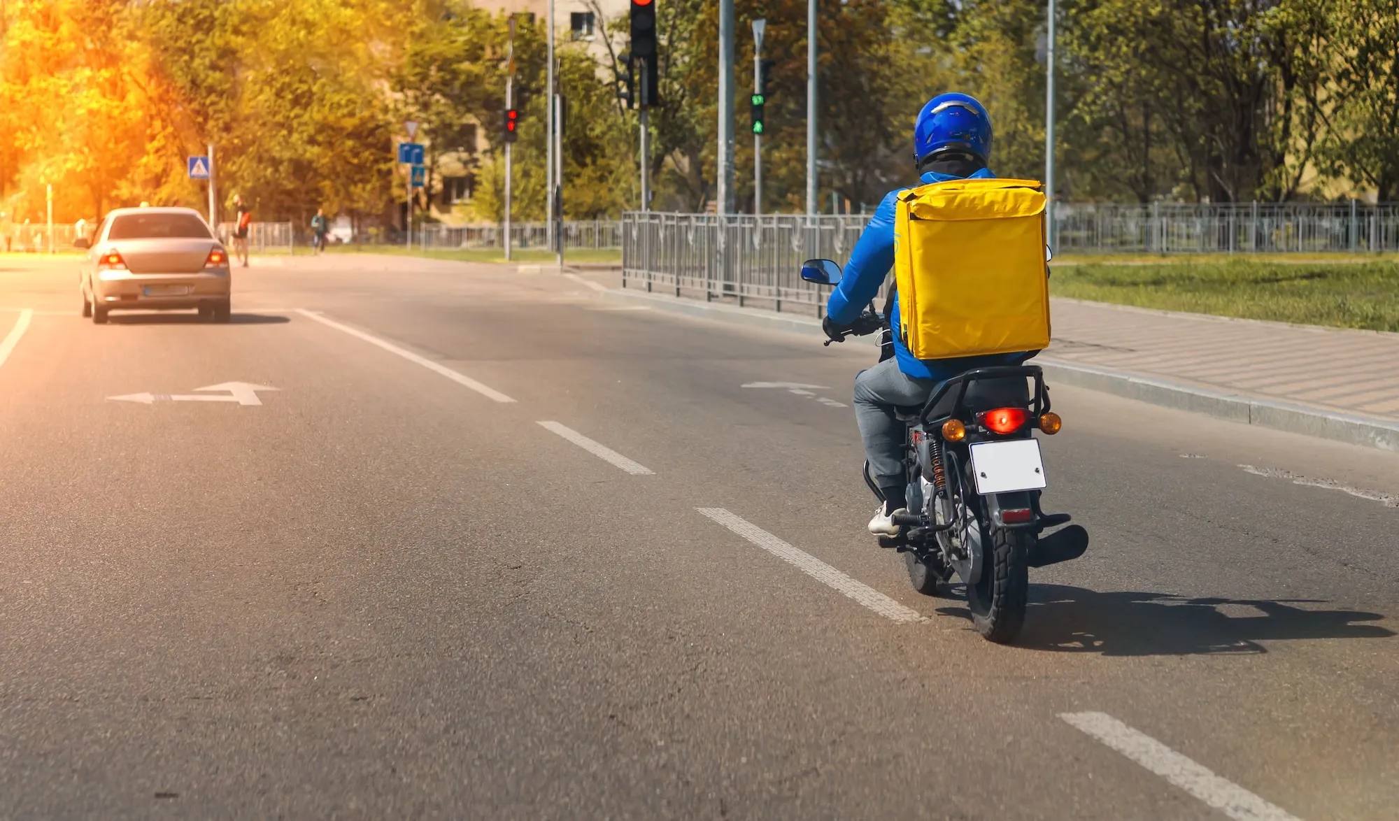 A person wearing a blue jacket and helmet rides a motorcycle with a large yellow delivery backpack down a city street in daylight, following a car toward a traffic light.