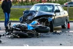A heavily damaged black car and a blue motorcycle are involved in a crash on a road, with debris scattered around. A person stands nearby, and the scene appears to be a serious accident.