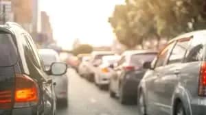 A line of cars is stuck in heavy traffic on a city street during daylight, sunlight reflecting off the vehicles and trees along the roadside—a common scene as drivers anticipate adjustments from California’s new traffic laws 2026.