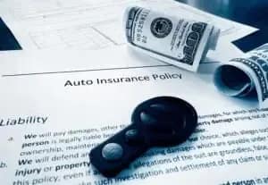 A car key, rolled-up cash, and paperwork labeled "Auto Insurance Policy" are shown on a table, symbolizing car insurance and financial planning.