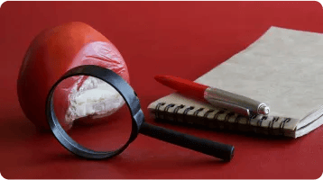 A bitten tomato, a magnifying glass, a red pen, and a spiral notebook are arranged on a red surface—capturing the careful investigation often required by a car accident attorney in personal injury cases.