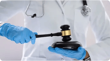 A person wearing a white lab coat, stethoscope, and blue gloves holds a gavel, symbolizing the intersection of healthcare and law or medical legal issues, such as those addressed by a personal injury attorney after car accidents.