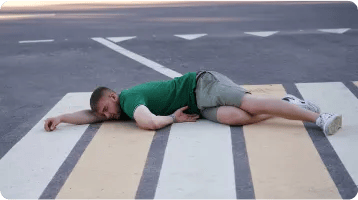 A man in a green shirt and shorts is lying on his side across a pedestrian crosswalk on an empty road, appearing tired or resting—possibly after one of the car accidents that often require the help of a car accident attorney.