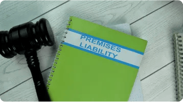 A green spiral notebook labeled "Premises Liability" lies on a white wooden surface next to a black judge's gavel and another partially visible notebook, highlighting the importance of personal injury knowledge for any personal injury attorney.