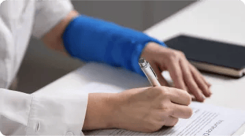 A person with a blue arm cast is sitting at a desk, filling out a form with a pen—perhaps seeking help from personal injury attorneys. A closed notebook rests nearby on the white surface.