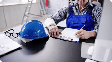 A person in blue overalls sits at a desk, writing on a clipboard. A blue hard hat and glasses are on the desk nearby, with office items in the background—suggesting careful documentation, much like a personal injury attorney preparing a case.