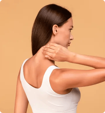 A woman with long brown hair, seen from the back, wears a white sleeveless top and touches her neck with her right hand as if experiencing personal injury discomfort, against a beige background.
