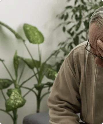 An elderly person wearing glasses and a beige sweater sits with their head resting on one hand, appearing stressed or deep in thought—perhaps considering consulting personal injury attorneys after car accidents. Green leafy plants are visible in the background.