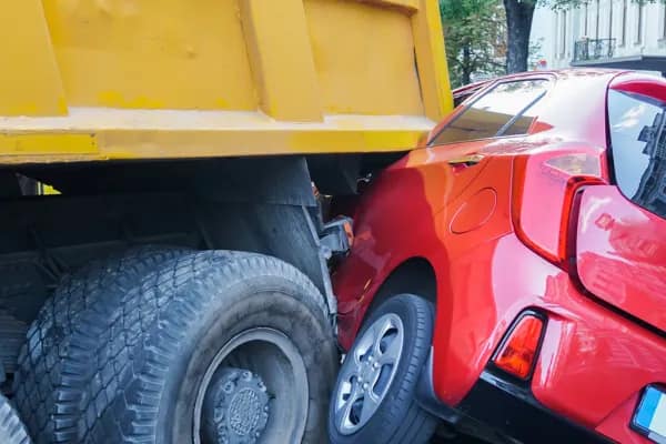 A red car is crushed under the back of a large yellow dump truck, its rear visibly damaged and wedged between the truck’s tires—a scene where a personal injury attorney could provide crucial legal assistance.