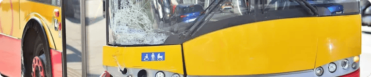 A yellow city bus with a shattered front windshield is parked near a red surface. The cracked glass on the driver's side hints at an accident—personal injury attorneys may be needed if anyone was hurt. Accessibility stickers are visible by the door.