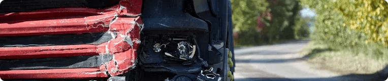 Close-up of a damaged red truck with a broken headlight and crumpled front corner, parked on the side of a rural road lined with trees—a stark reminder of why consulting a car accident attorney after car accidents is important.