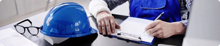 A person with a bandaged arm is filling out paperwork on a clipboard at a desk, with a blue safety helmet and eyeglasses nearby—possibly documenting injuries for a personal injury claim after car accidents.