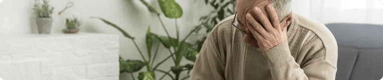 An elderly man sits indoors on a couch, wearing a beige sweater and glasses, resting his head in one hand, appearing stressed or deep in thought—perhaps after dealing with personal injury from car accidents. Green plants and white decor fill the background.