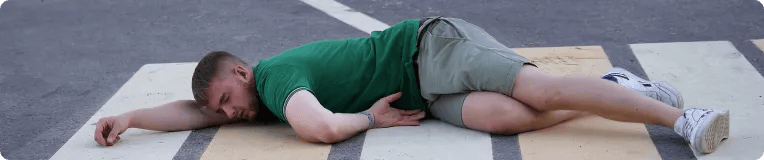 A man wearing a green shirt and gray shorts is lying face down on a pedestrian crosswalk on an asphalt road, a scene that often requires the assistance of personal injury attorneys.
