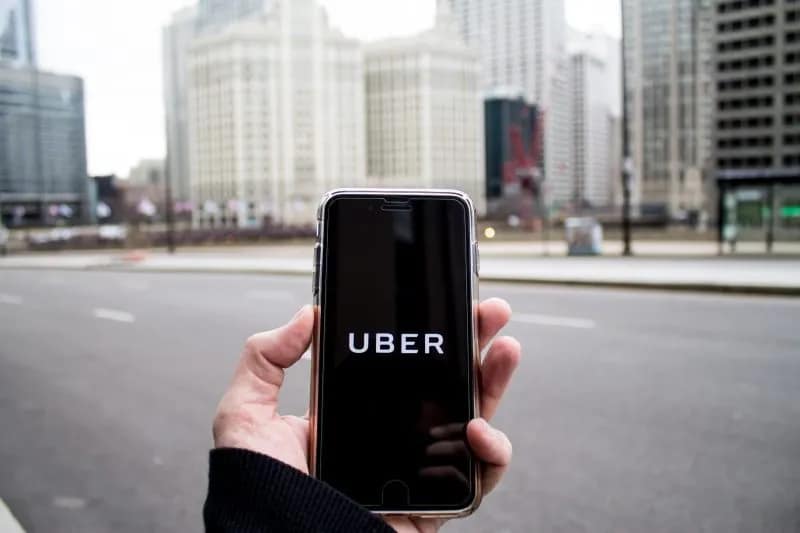A person holds a smartphone displaying the Uber app logo on the screen, with tall city buildings and an empty street in the background—an important scene for lawyers handling car accidents in urban areas.