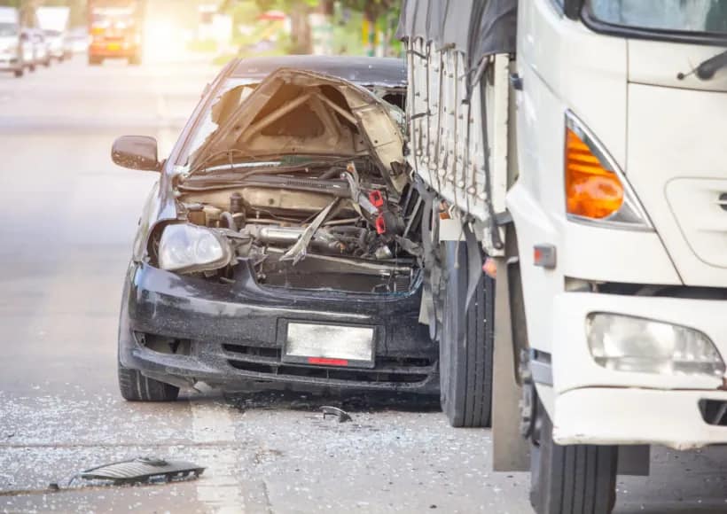 A black car with a severely smashed front end is wedged under the back of a large white truck on a road. Broken glass and car parts are scattered on the ground, underscoring the need for a personal injury lawyer after such an accident.