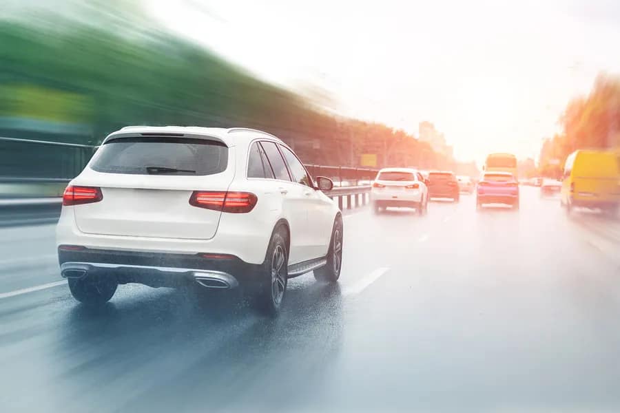 A white SUV drives on a wet, multi-lane road with blurred cars and trees, suggesting motion—an important scene for lawyers specializing in car accidents, as busy roads like this often lead to legal cases.