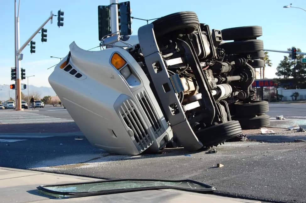 A large white truck is overturned on its side in the middle of a city intersection, with debris and a detached windshield nearby—an incident where a personal injury attorney may be needed. Traffic lights and buildings are seen in the background.