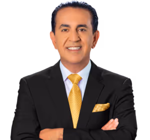 Un hombre con un traje negro, camisa blanca y corbata amarilla con patrón está de pie con los brazos cruzados, sonriendo a la cámara contra un fondo claro. Tiene el cabello oscuro peinado hacia atrás y lleva un pañuelo amarillo a juego.