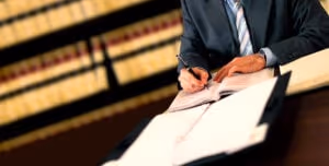 A person in a suit writes in an open book at a desk, with a binder and legal bookshelves in the background, suggesting a personal injury attorney working on car accidents cases in a professional setting.