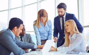Cinco abogados vestidos profesionalmente se reúnen alrededor de una mesa en una oficina, discutiendo algo en una laptop. Parecen estar colaborando en casos de abogados de lesiones personales, con papeles y vasos de agua sobre la mesa. Grandes ventanas están en el fondo.