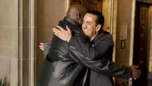 Dos hombres, uno con una chaqueta de cuero negra y el otro con un traje oscuro, están sonriendo y abrazándose cálidamente en un entorno interior ornamentado—quizás celebrando una gran victoria como abogados de lesiones personales.