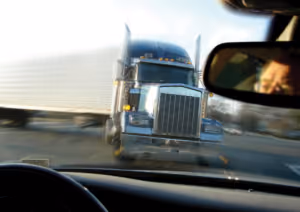 Vista desde dentro de un automóvil mientras un gran camión semi se acerca de frente a alta velocidad, evocando el peligro de los accidentes automovilísticos. Se refleja una persona en el espejo retrovisor, destacando momentos que pueden requerir un abogado de lesiones personales.