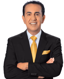 A man in a black suit with a white shirt and a yellow patterned tie stands with arms crossed, smiling at the camera. He has dark hair neatly styled and a matching yellow pocket square. The background is plain and light-colored.