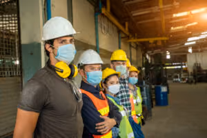 Un grupo de trabajadores de fábrica con cascos, chalecos de seguridad y mascarillas de protección están uno al lado del otro dentro de un almacén industrial, mirando en la misma dirección—muy parecido a un equipo de abogados de lesiones personales listos para abogar por la seguridad en el lugar de trabajo.