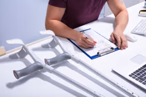 Una persona sentada en un escritorio llena un formulario en un portapapeles, con muletas descansando sobre la mesa cerca—sugiriendo que pueden estar lidiando con una lesión personal—junto a una laptop y un teclado.