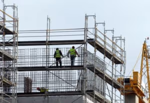Two construction workers in high-visibility vests stand on scaffolding at a building site, surrounded by metal bars and framework, with a yellow crane in the background—reminding us why safety matters, just like lawyers do after car accidents.
