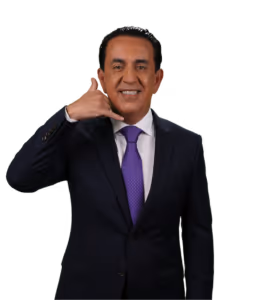 A man in a dark suit and purple tie smiles while making a "call me" gesture with his hand, holding his thumb and pinky finger to his face. The background is plain white.