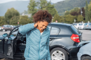 A woman stands next to her car in a parking lot, holding her neck and wincing in pain after a car accident. Both vehicles show signs of collision, highlighting the personal injury risks often faced in car accidents.