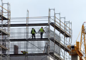 Two construction workers in high-visibility vests stand on scaffolding at a building site, surrounded by metal bars and framework, with a yellow crane in the background—reminding us why safety matters, just like lawyers do after car accidents.