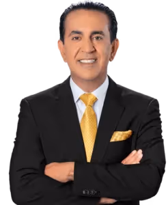 A man in a black suit with a white shirt and a yellow patterned tie stands with arms crossed, smiling at the camera. He has dark hair neatly styled and a matching yellow pocket square. The background is plain and light-colored.