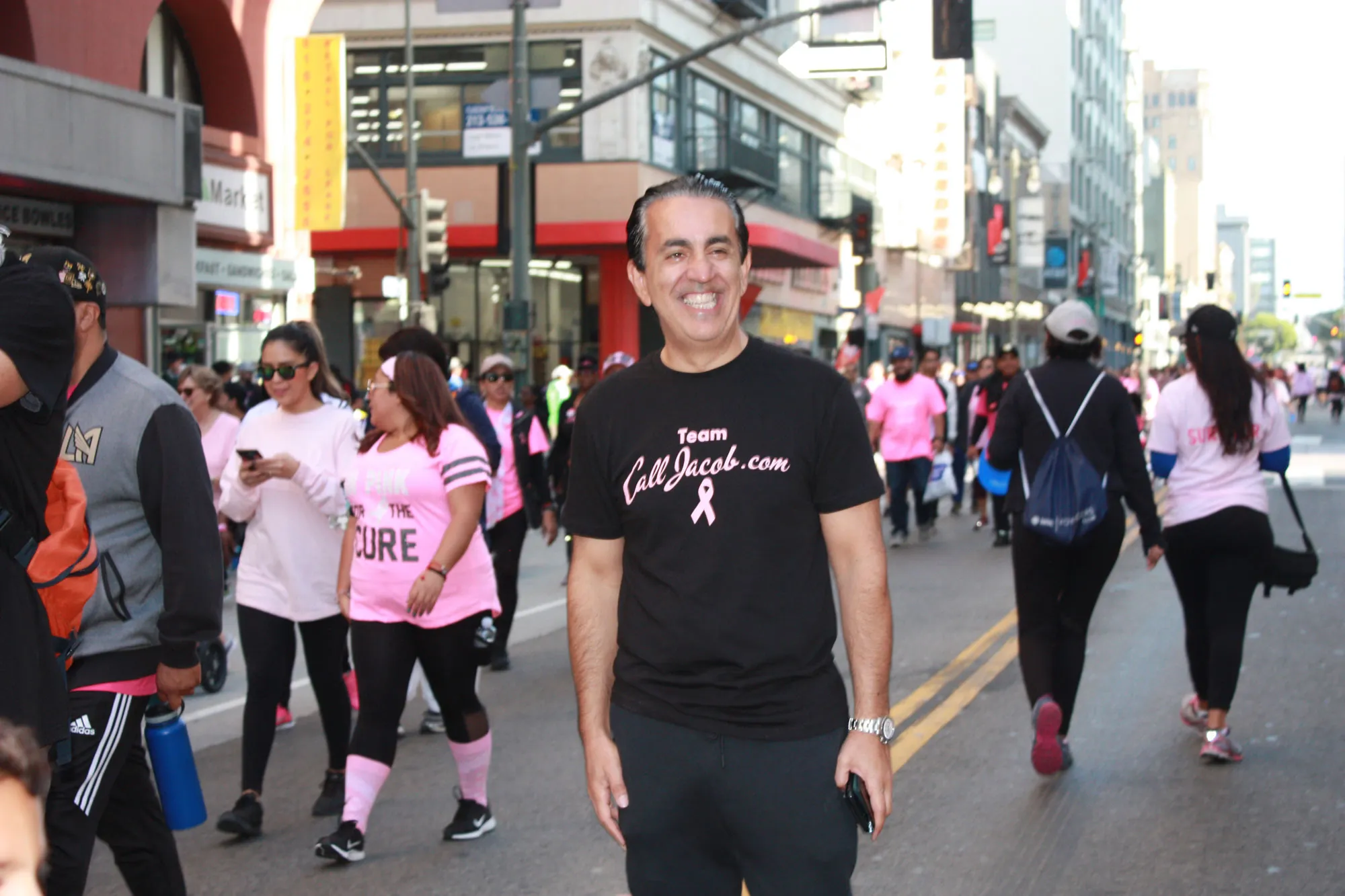 A man wearing a black "Team ValJacobs.com" shirt and smiling stands in the middle of a city street during a breast cancer awareness walk, surrounded by people in pink shirts, showing support as a personal injury attorney.