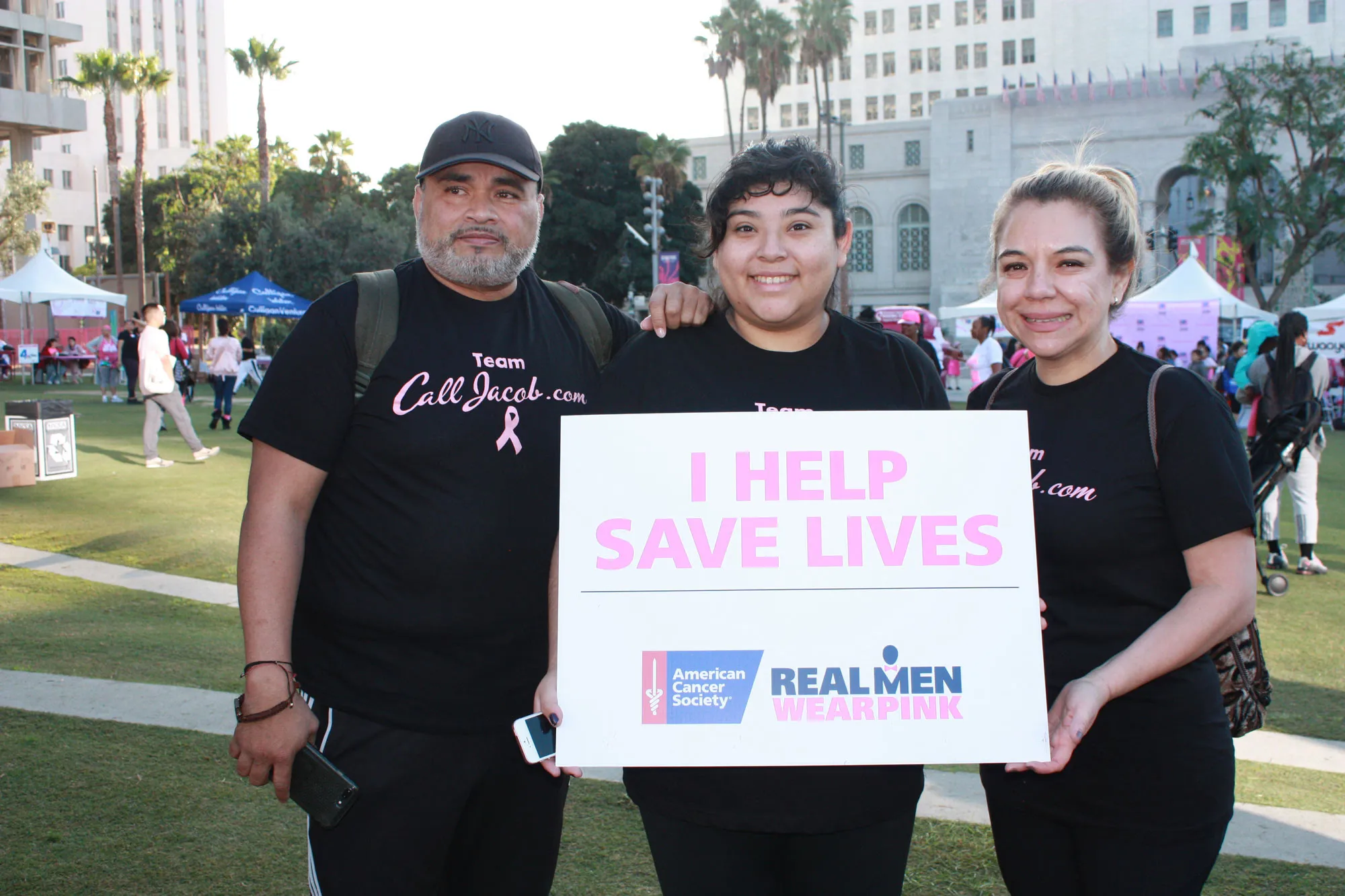 Three people smile while holding a sign that says "I HELP SAVE LIVES" at an outdoor event. Wearing black shirts with a pink ribbon, they support the American Cancer Society's "Real Men Wear Pink" campaign—showing even lawyers care beyond car accidents.