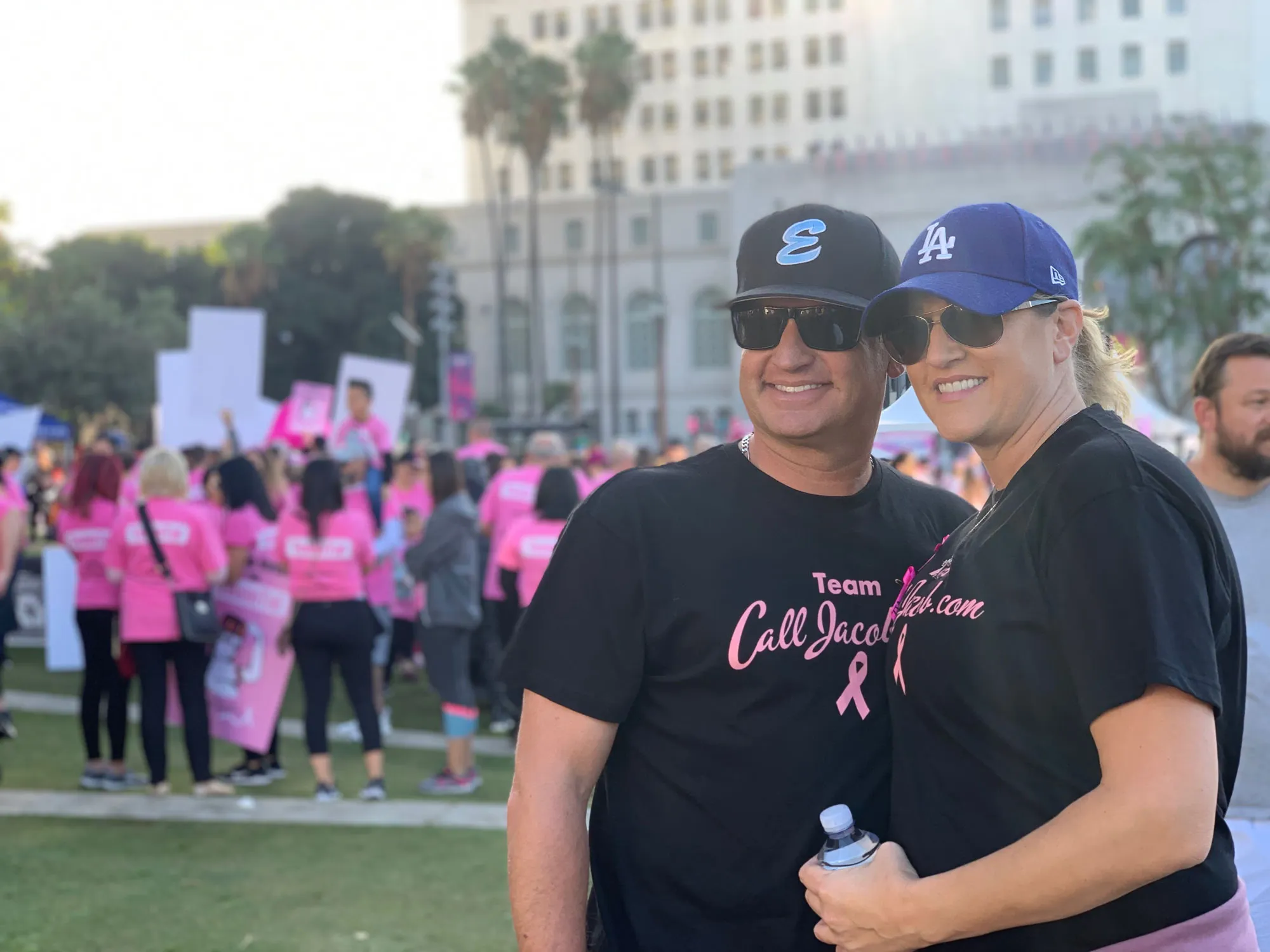 Two people wearing black shirts with pink writing and baseball caps stand together, smiling at an outdoor event. In the background, a crowd in pink shirts supports a breast cancer awareness walk or run—important for causes like car accidents and personal injury.
