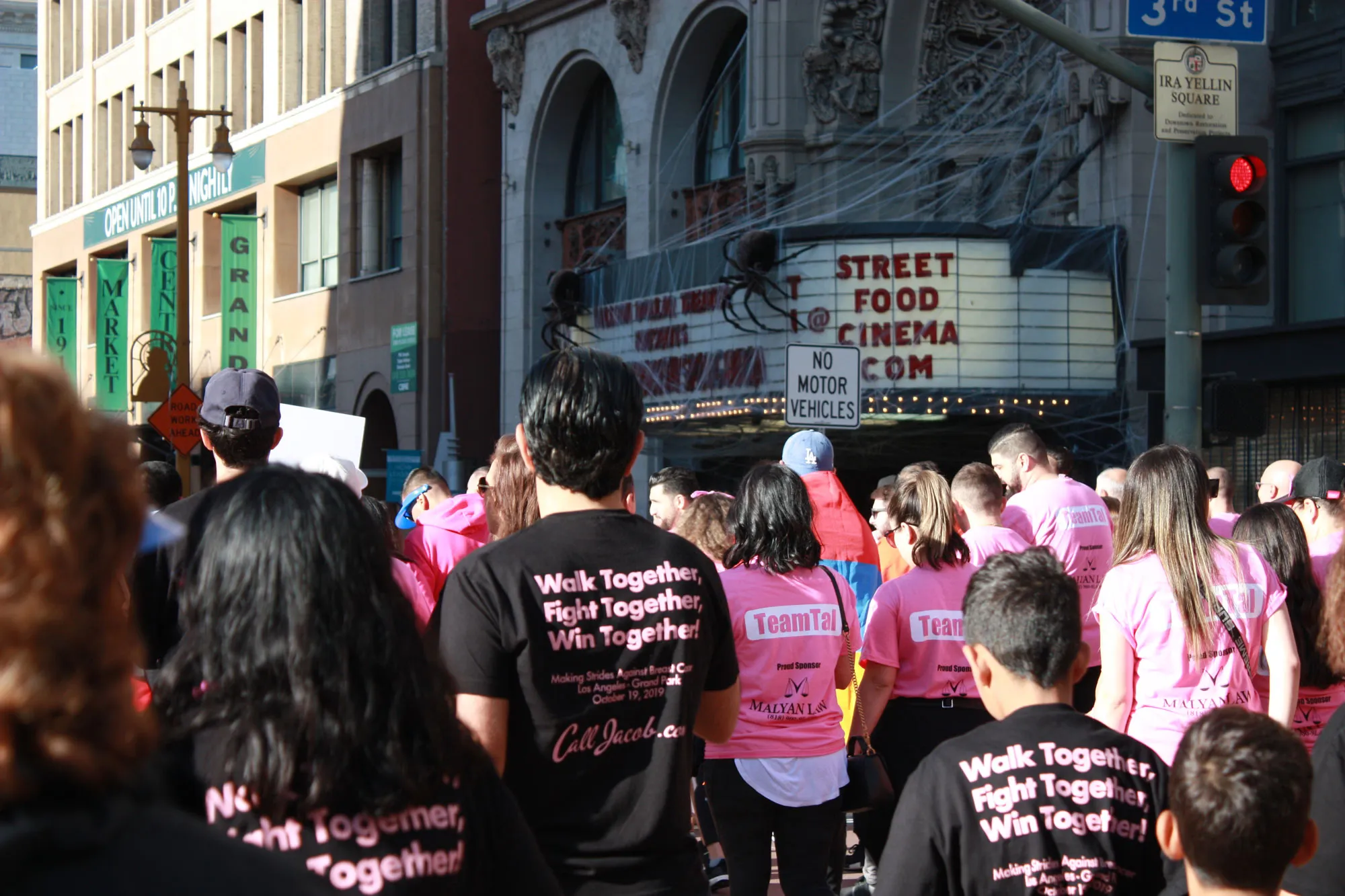 A crowd of people wearing pink and black shirts with motivational slogans participates in a walk event on a city street, raising awareness about car accidents. A theater marquee in the background reads "STREET FOOD CINEMA.COM.