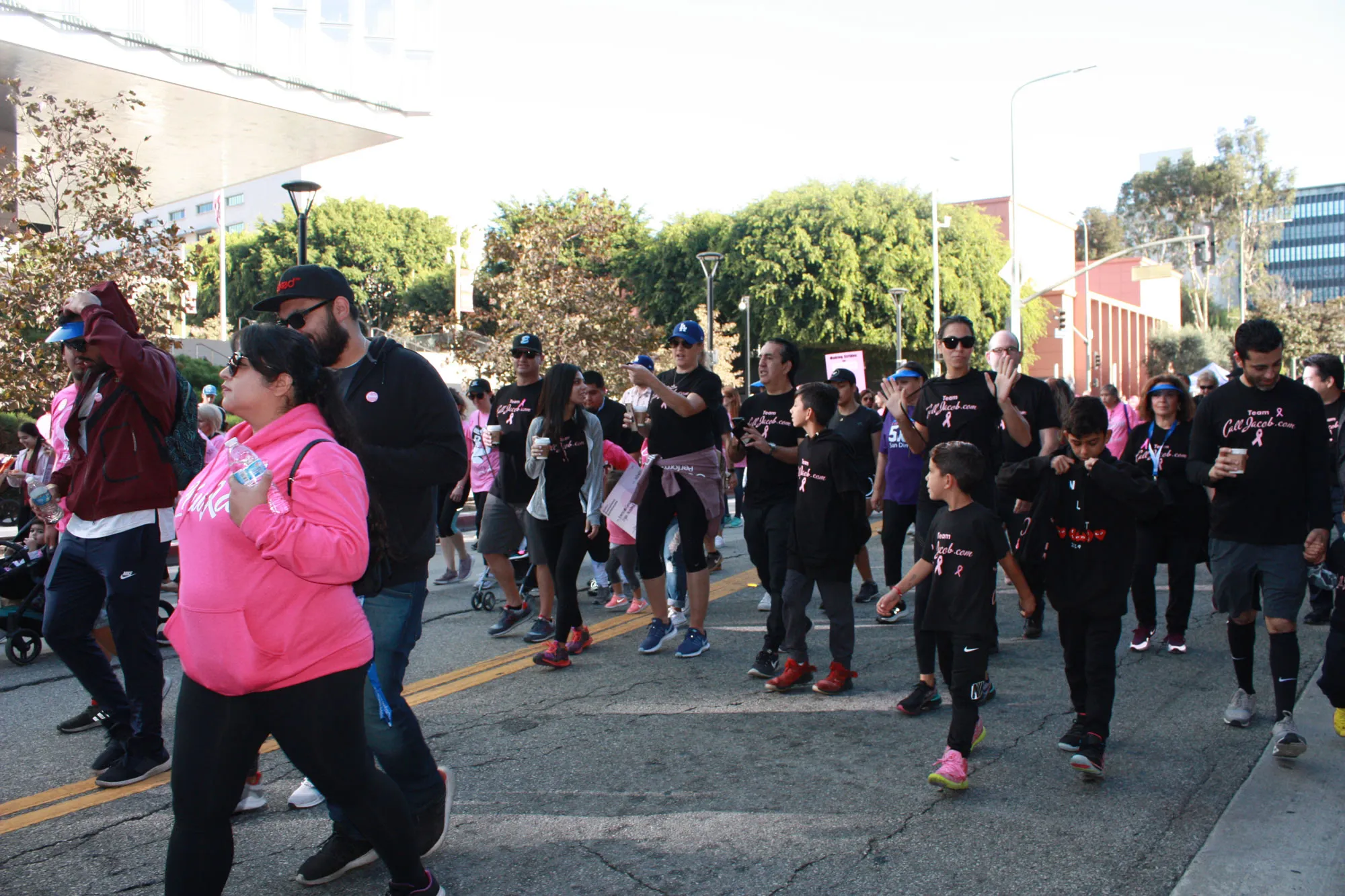 A group of people, many wearing matching black shirts with pink writing and pink ribbons, walk together during a street event, possibly a charity walk or fundraiser. Among them are personal injury attorneys supporting the cause.