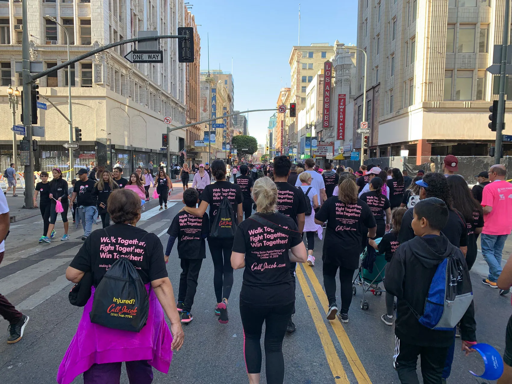 A crowd of people in matching black shirts with pink writing participate in a city street walk event for personal injury awareness, surrounded by tall buildings and street signs. Many shirts read, “Walk Together, Fight Together, Win Together!”.