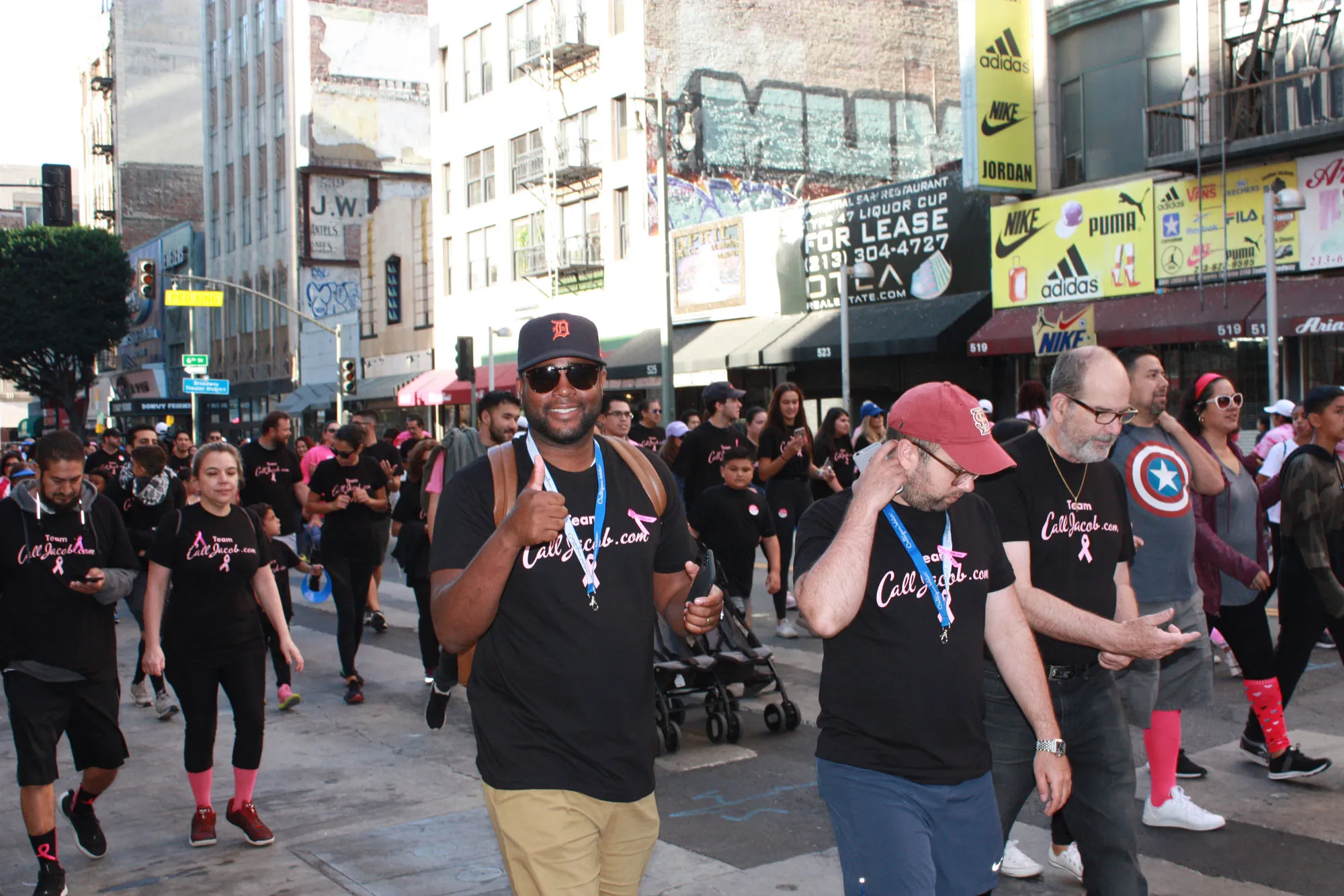 A group of people wearing matching black "CalifaCare" shirts and pink ribbons walk together on a city street, raising awareness for personal injury support during a community event. Some participants are smiling, and the street is lined with shops and buildings.