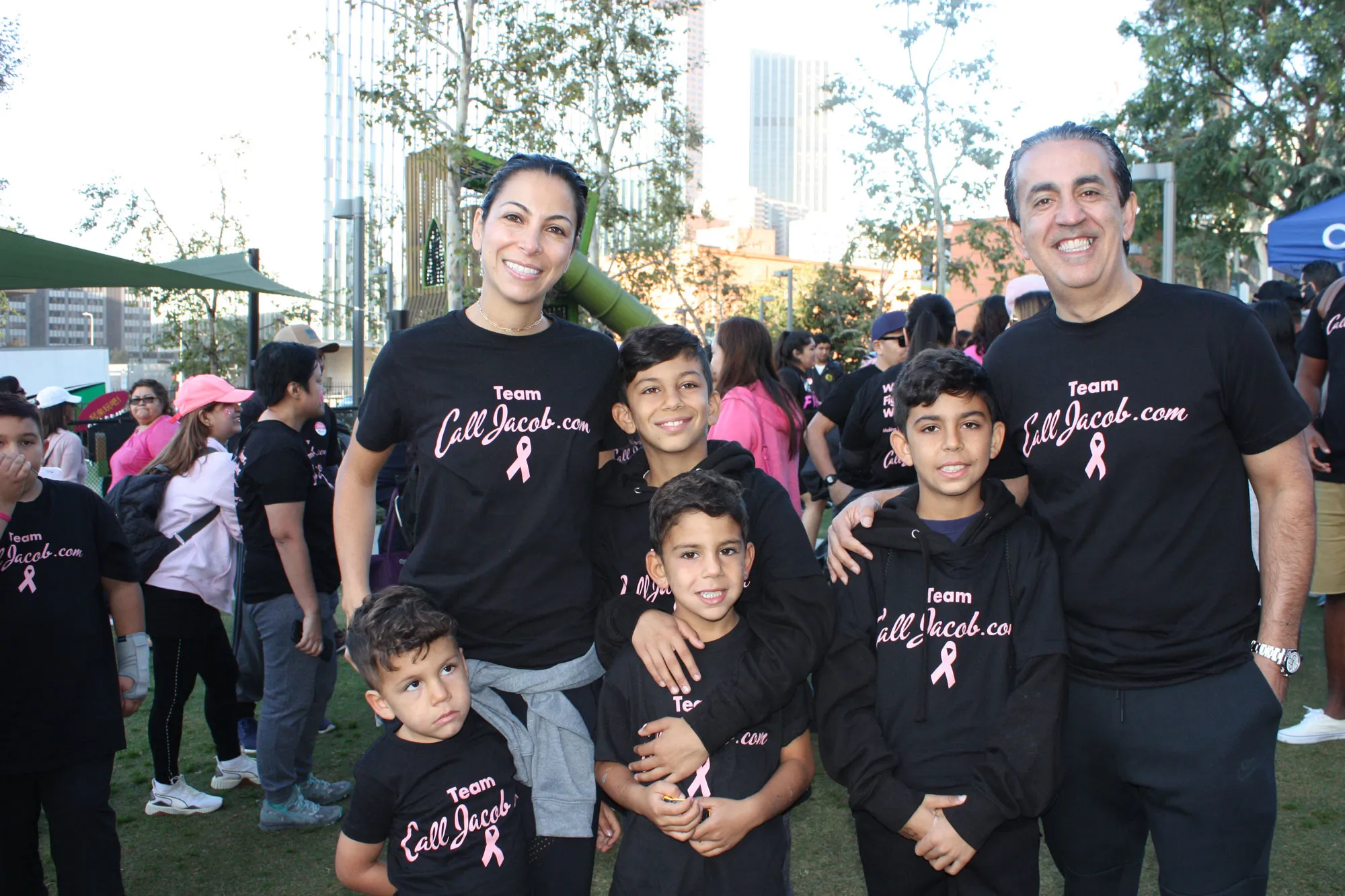 A smiling family and children, all in matching black “Team liljacob.com” shirts with a pink ribbon, pose together at an outdoor charity event to raise awareness for car accidents. Other participants and trees fill the background.