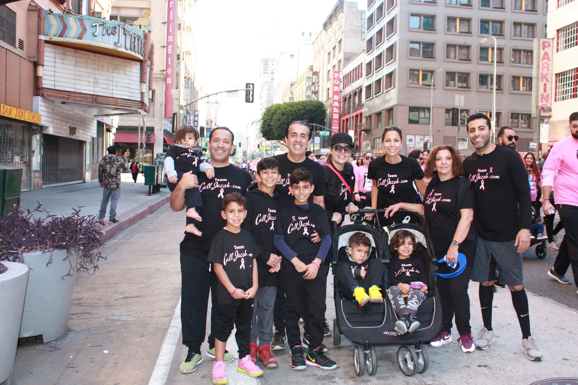 A group of people, including adults and children, smile and pose on a city street wearing matching black shirts with pink ribbons for a charity walk. Among them are lawyers raising awareness for car accidents and personal injury attorney services. Some kids are in a stroller.