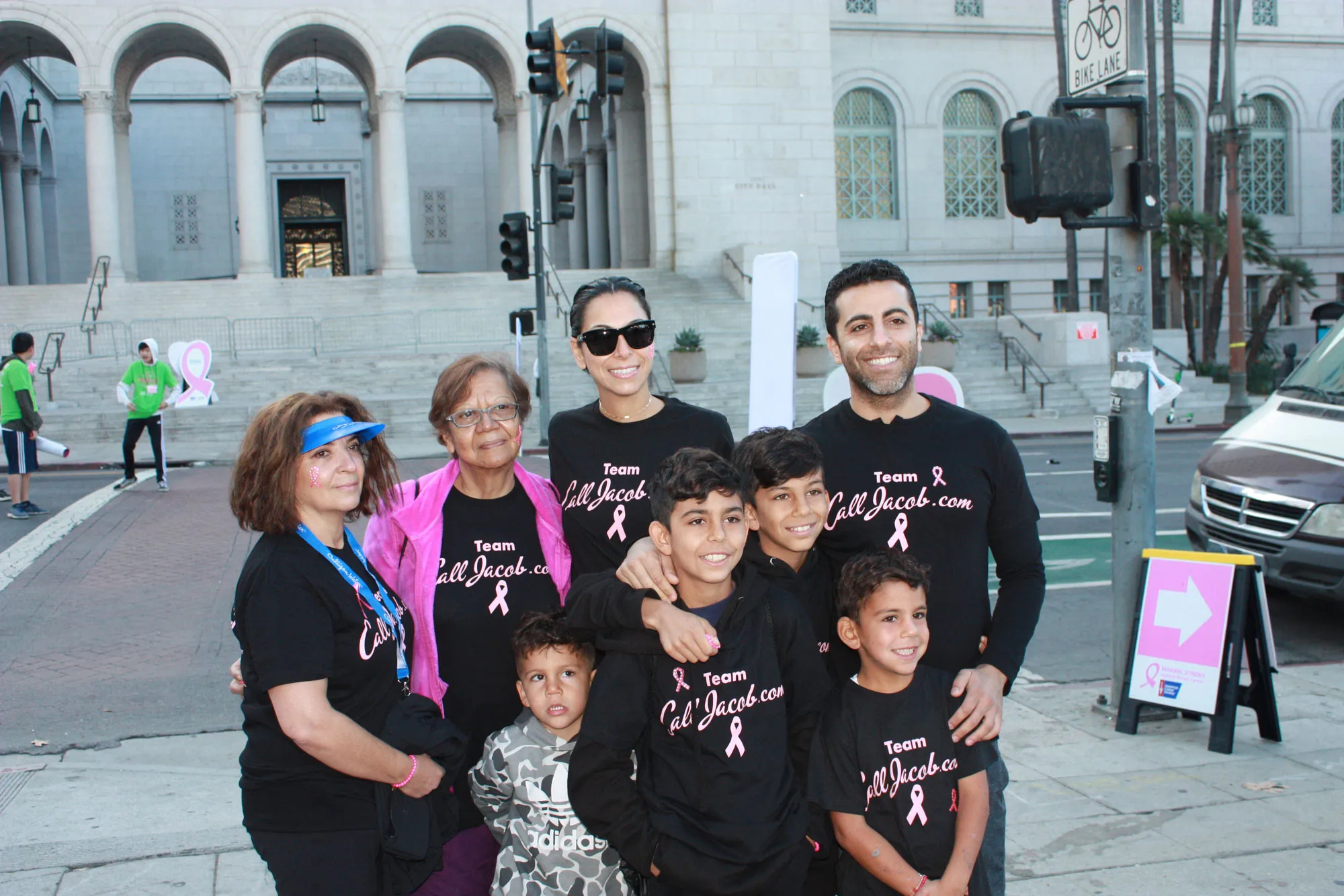 A group of people, adults and children, wearing matching "Team Lil' Jackie" shirts, smile together at a breast cancer awareness event on a city street—just steps away from where a local personal injury attorney’s office is visible in the background.