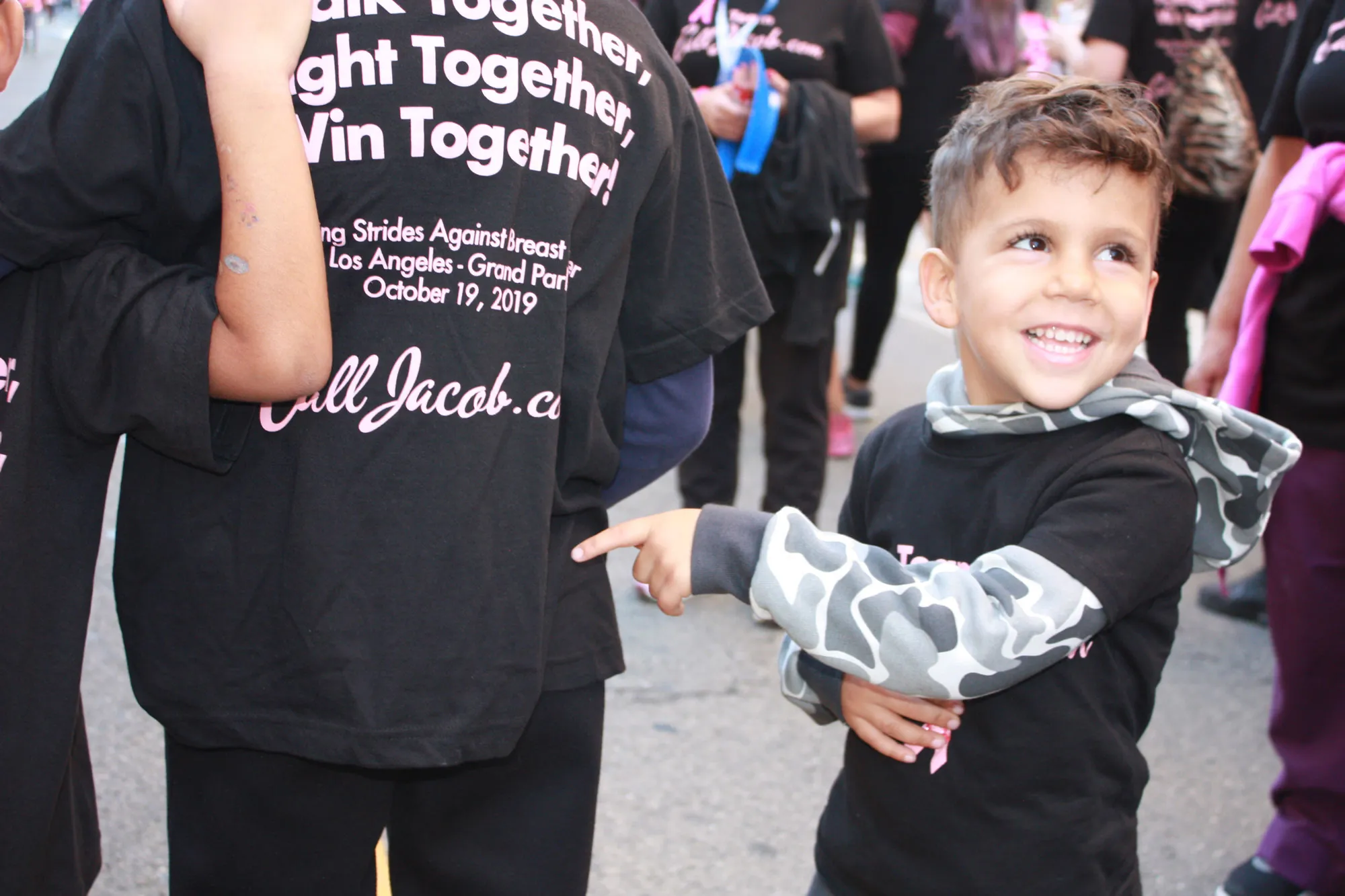 A young boy in a gray camouflage hoodie smiles and points at an older child’s t-shirt at a breast cancer awareness event, surrounded by people in black event shirts, including lawyers supporting the cause.