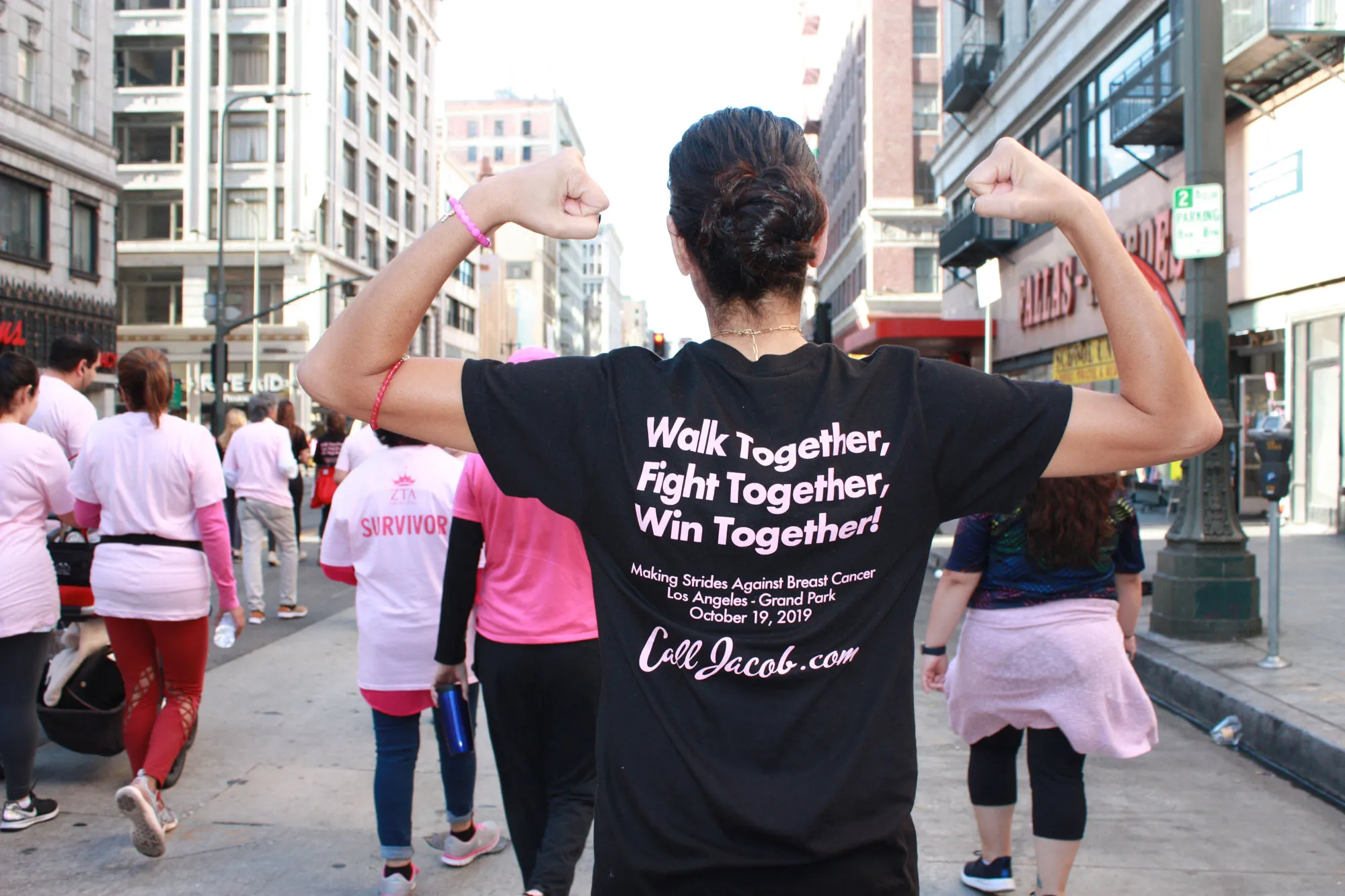 A person faces away from the camera, flexing their arms on a city street during a breast cancer awareness walk. Their shirt reads, "Walk Together, Fight Together, Win Together!" Lawyers in pink shirts walk ahead.