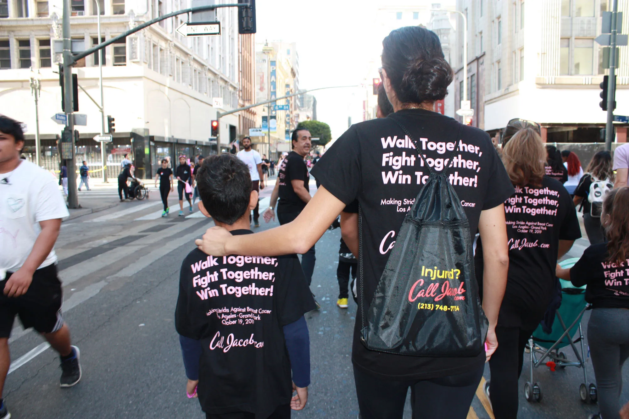 A woman with her arm around a child walks in a city street alongside others during an event. They wear matching black shirts with “Walk Together, Fight Together, Win Together!” printed on the back to support car accident attorney awareness.